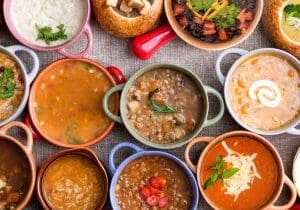 Variety of Garnished Soups in Colorful Bowls High Angle View of Various Comforting and Savory Gourmet Soups Served in Bread Bowls and Handled Dishes and Topped with Variety of Garnishes on Table Surface with Gray Tablecloth